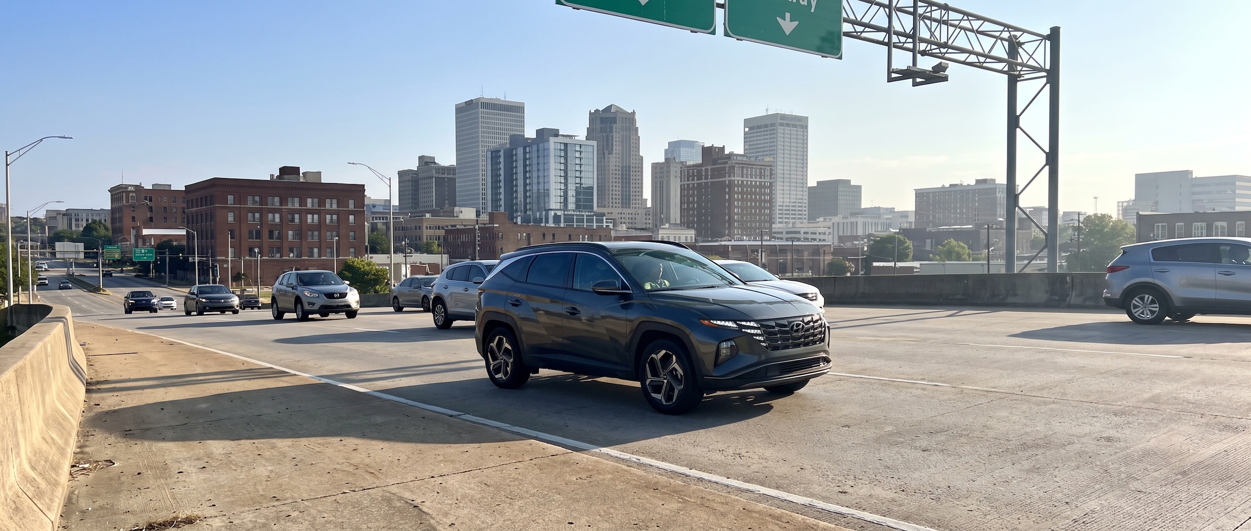 A 2026 Hyundai Tucson Hybrid in a modern charcoal finish exiting a downtown Birmingham interstate ramp with the city skyline visible in the background on a bright summer morning in 2026, late-July light, green highway signs visible overhead.