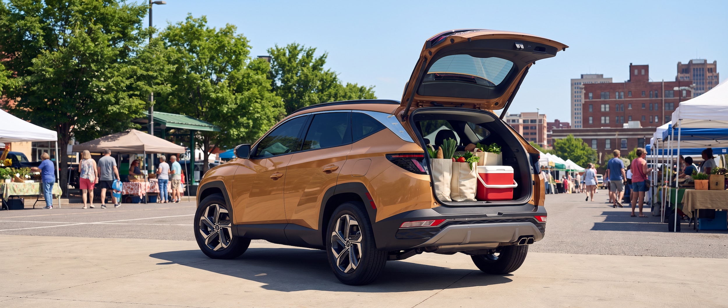 A 2026 Hyundai Tucson in a warm metallic color, liftgate open, parked at an outdoor farmers market on a bright Alabama summer morning, cargo area loaded with canvas produce bags and a small cooler, Birmingham urban backdrop with greenery