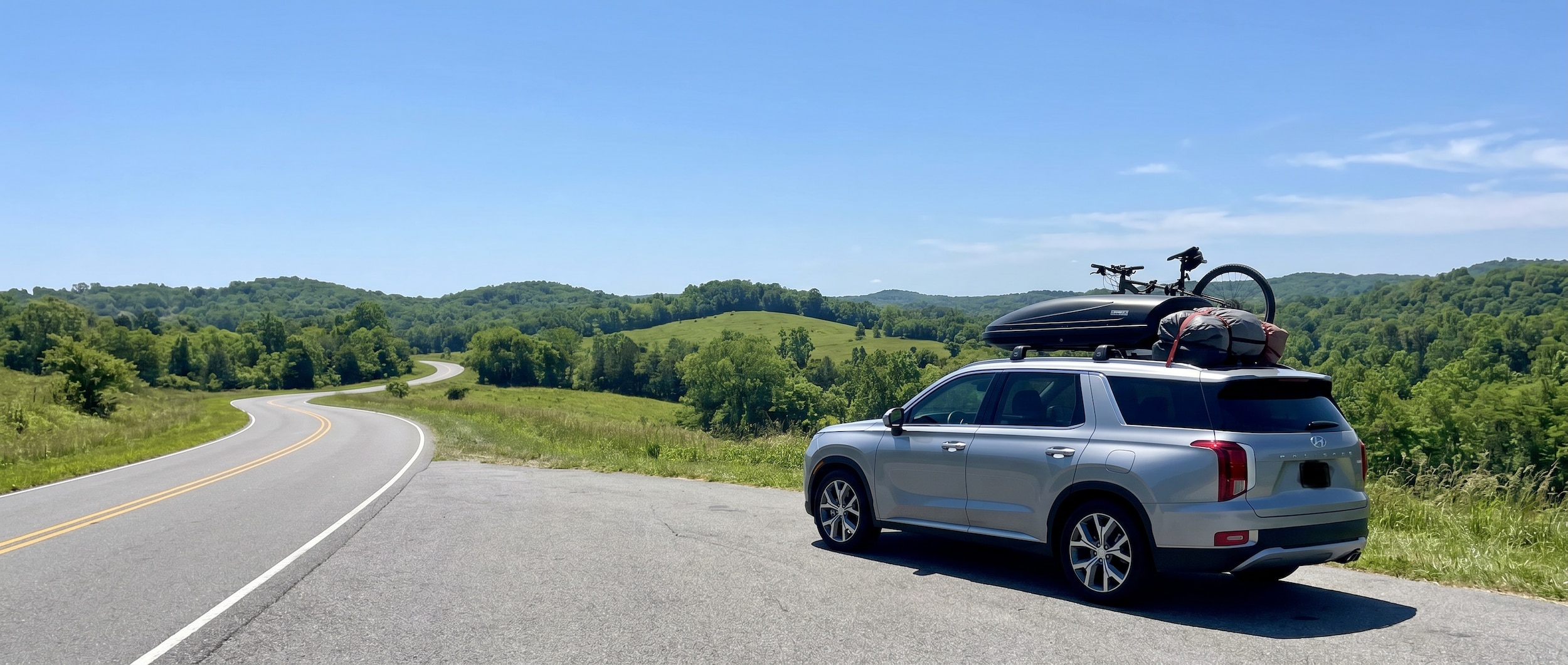 A silver Hyundai Palisade loaded with luggage, parked at a scenic overlook along an Alabama highway on a bright summer day, American flags visible on the dashboard, blue sky and lush green hills in the background.