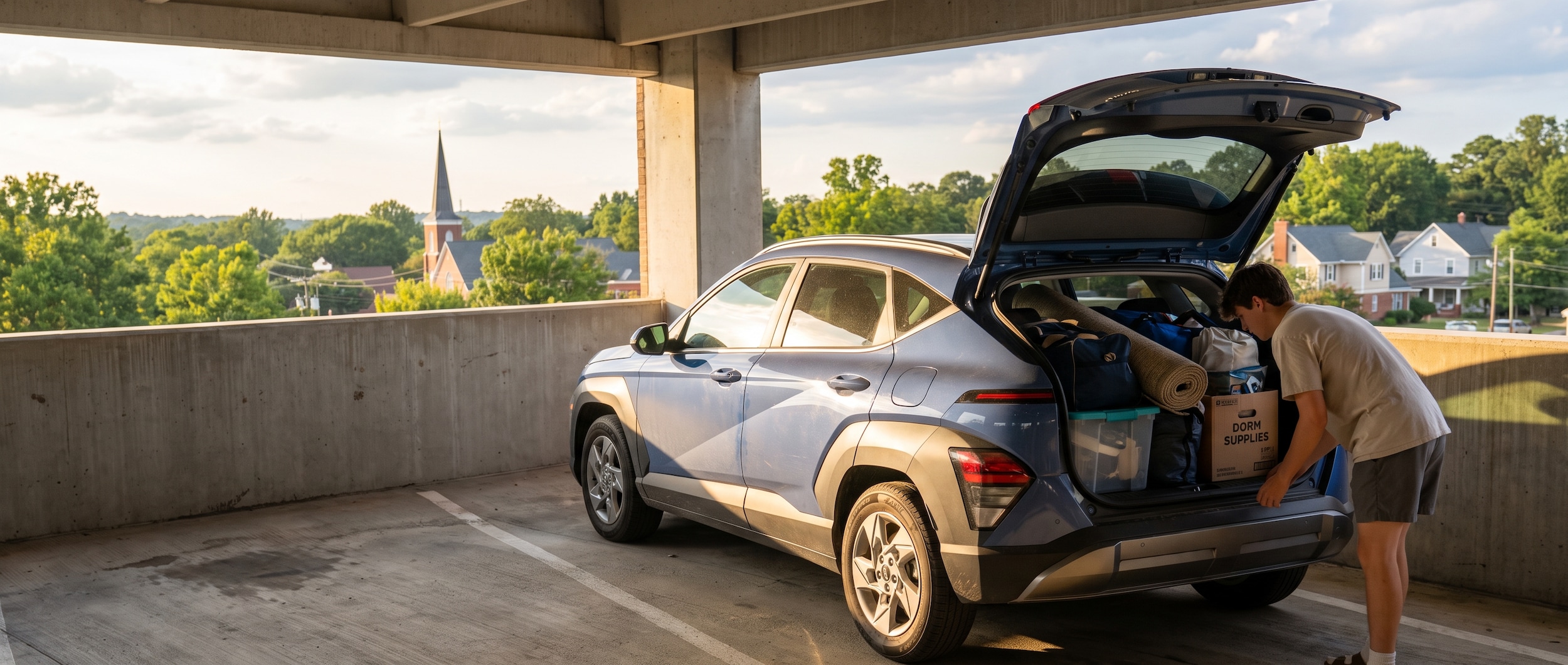 A 2026 Hyundai Kona in a compact campus parking deck, back hatch open and loaded with dorm gear, warm Alabama summer light, Homewood suburban backdrop visible through the open deck wall