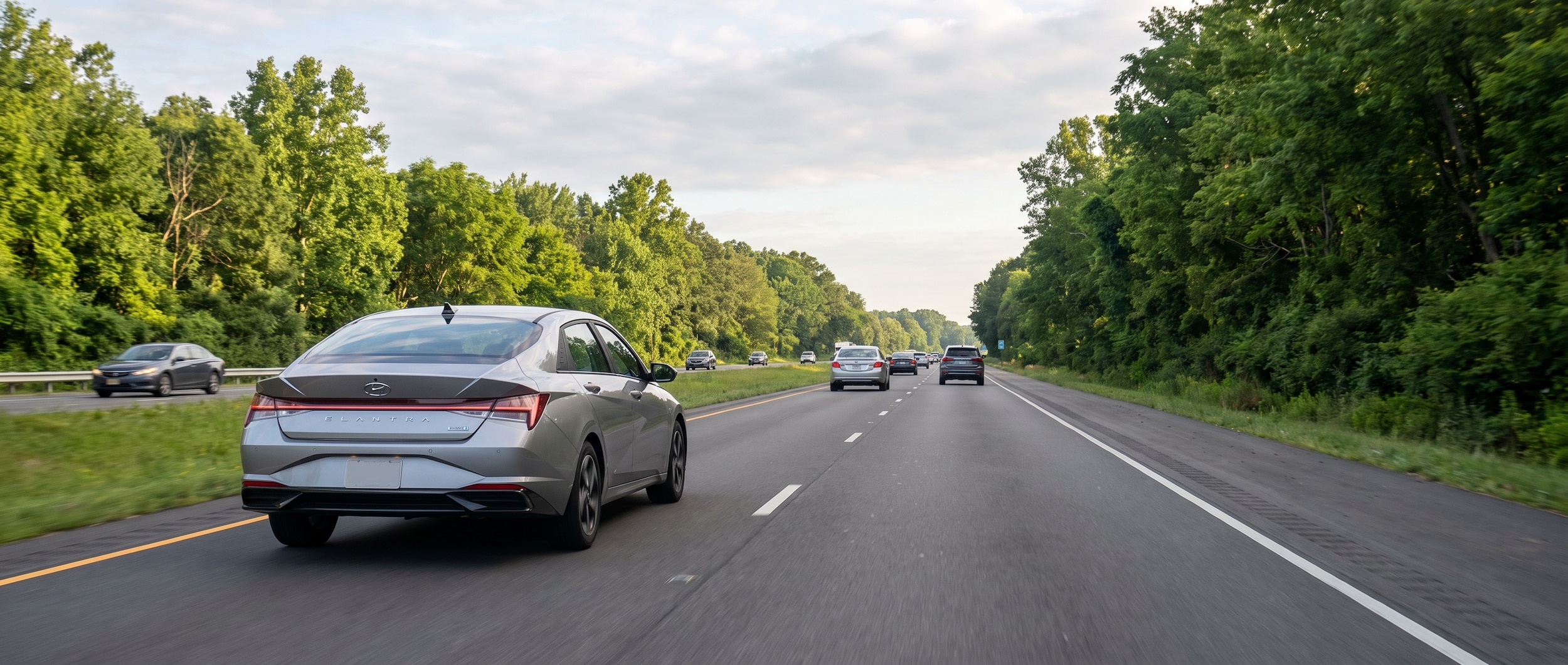 A 2026 Hyundai Elantra Hybrid in silver or blue metallic driving on a wide multi-lane suburban highway with green Alabama summer foliage on both sides, soft morning light, light commuter traffic ahead suggesting the US-280 corridor near Birmingham