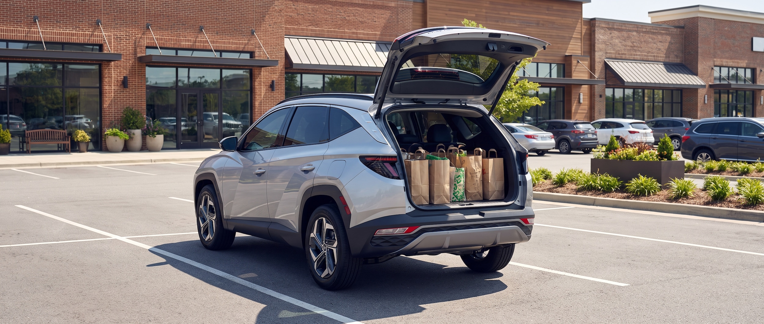 A 2026 Hyundai Tucson Hybrid in Shimmering Silver parked in a lifestyle shopping center lot on a bright Alabama summer day, rear hatch open showing cargo area loaded with shopping bags, storefronts visible in background