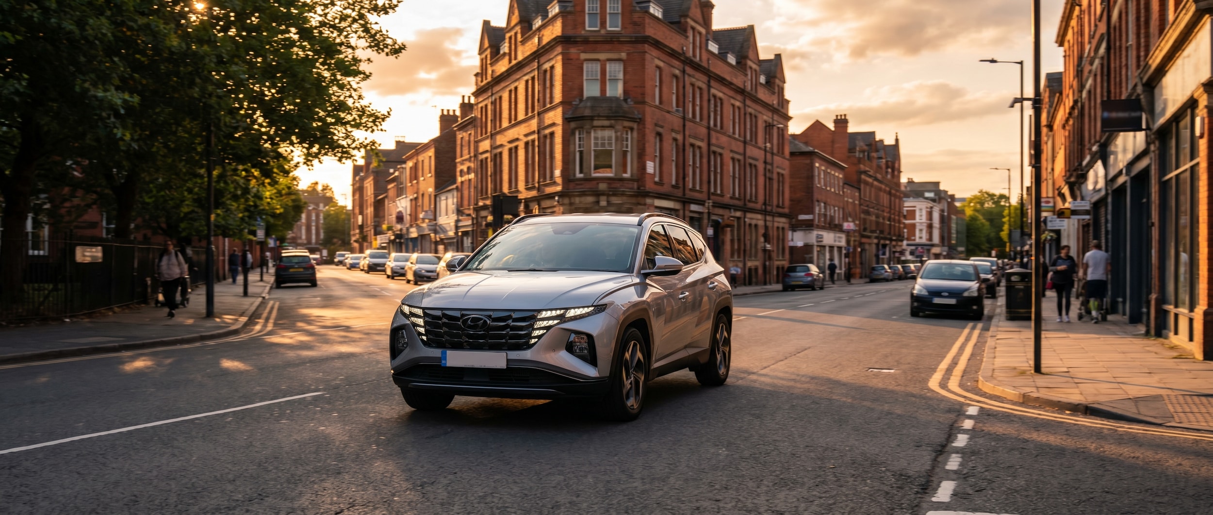A silver Hyundai Tucson Hybrid driving through an urban Birmingham street during golden hour, with city buildings visible in the background and tree-lined roads ahead, conveying a smooth and efficient daily commute.