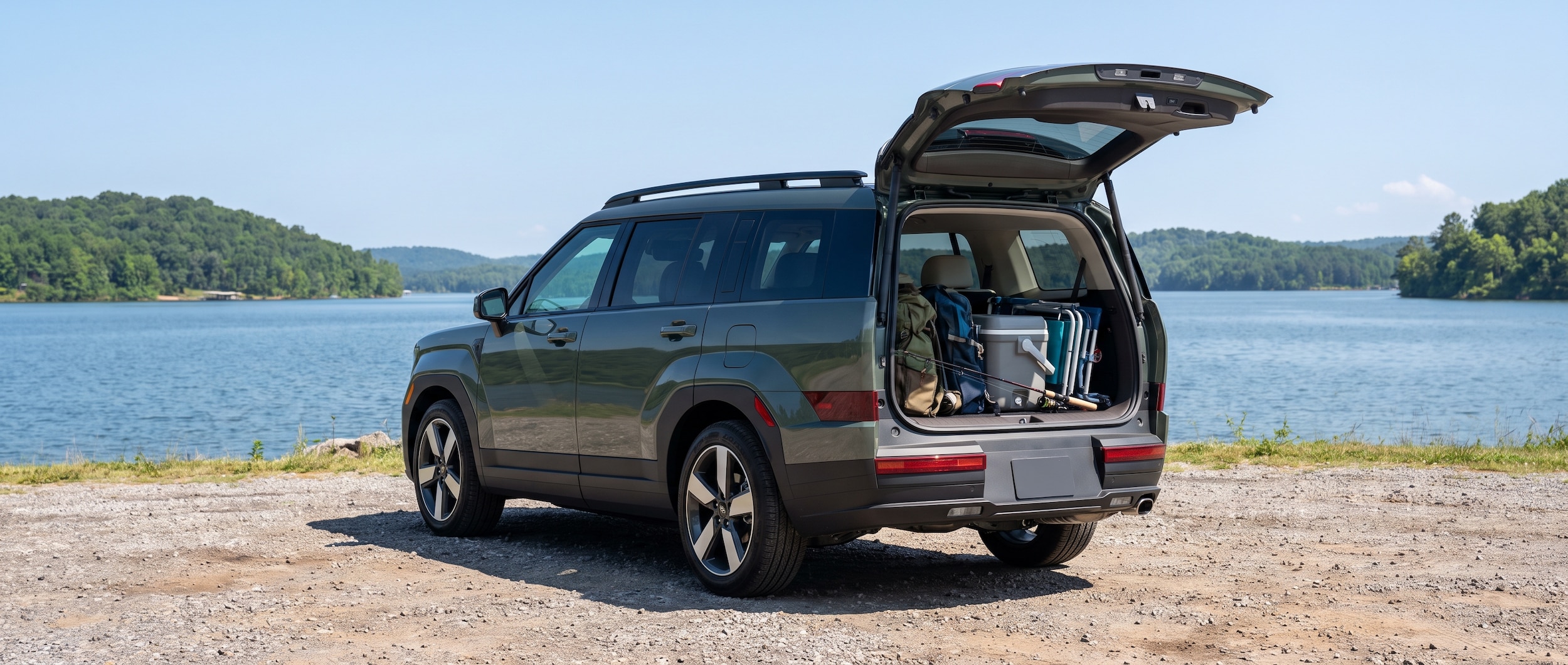 A 2026 Hyundai Santa Fe Hybrid in a lakeside setting at Lake Guntersville State Park, Alabama, with clear summer skies and the lake visible in the background -- the SUV is parked on a gravel pull-off, rear liftgate open, gear visible inside