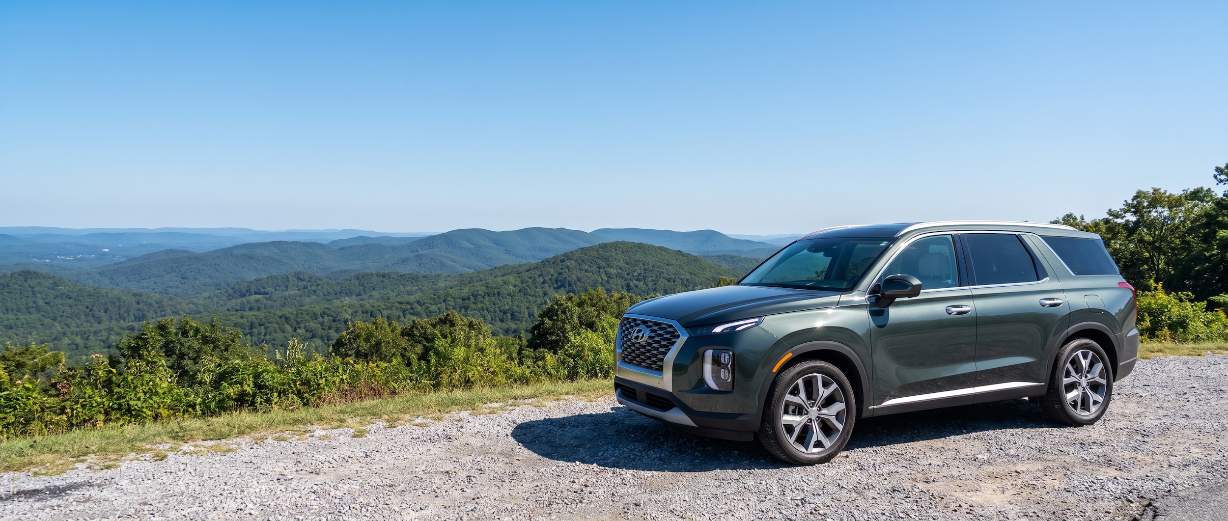 A scenic mountain overlook at Cheaha State Park with a modern Hyundai SUV parked at a pulloff, tree-covered ridgelines stretching into the distance under a clear blue Alabama sky