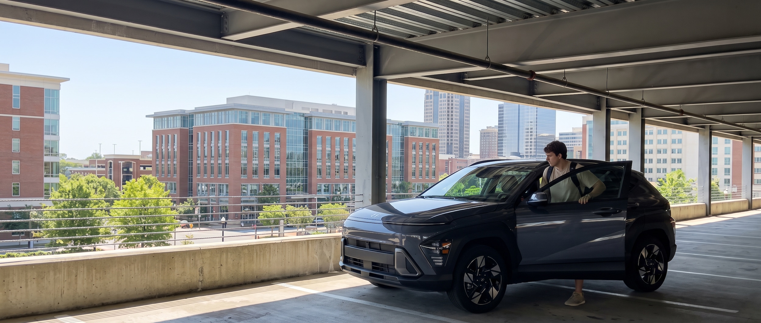 A 2026 Hyundai Kona in a compact urban parking deck near a university campus, midday summer light, driver-side door open, modern city buildings visible in the background suggesting a Birmingham downtown setting