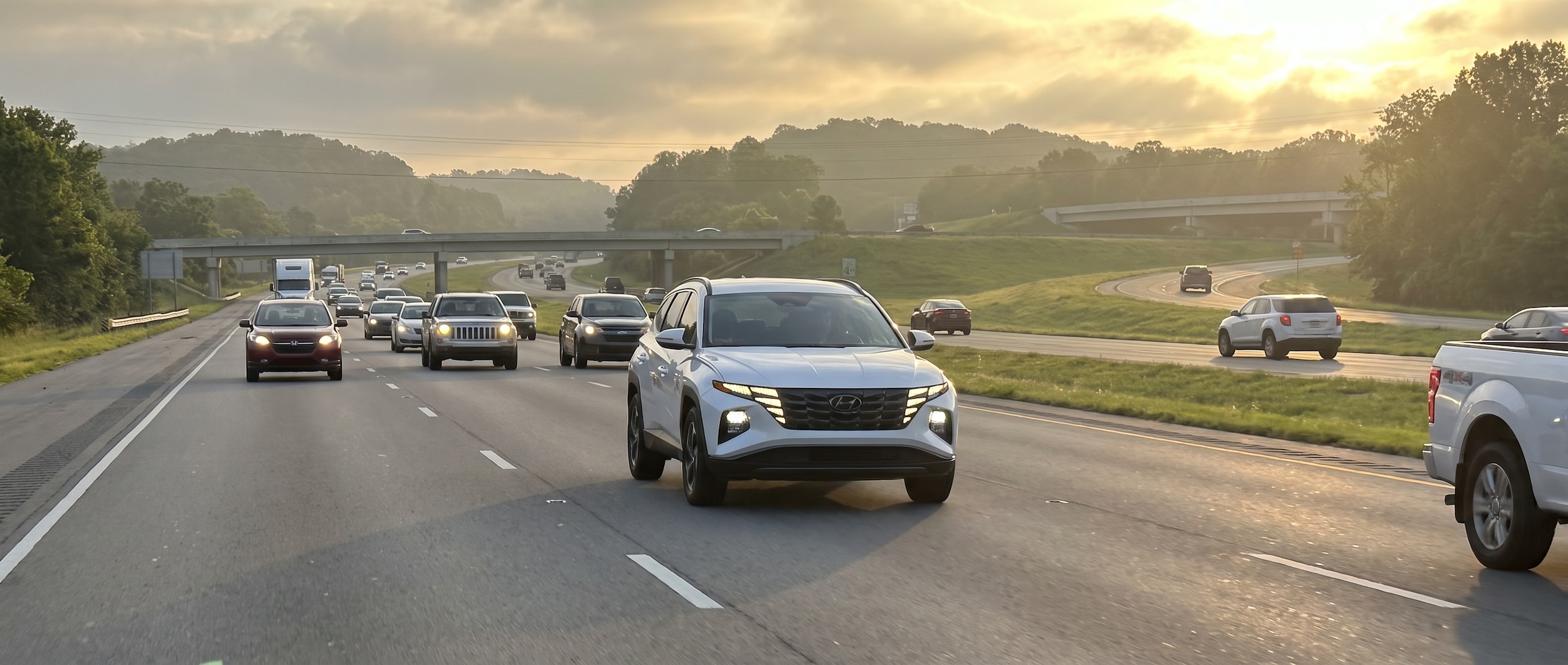 A 2026 Hyundai Tucson Hybrid in Serenity White Pearl navigating a multi-lane suburban highway corridor at morning rush hour, green hills and overpass structures visible, warm Alabama summer light