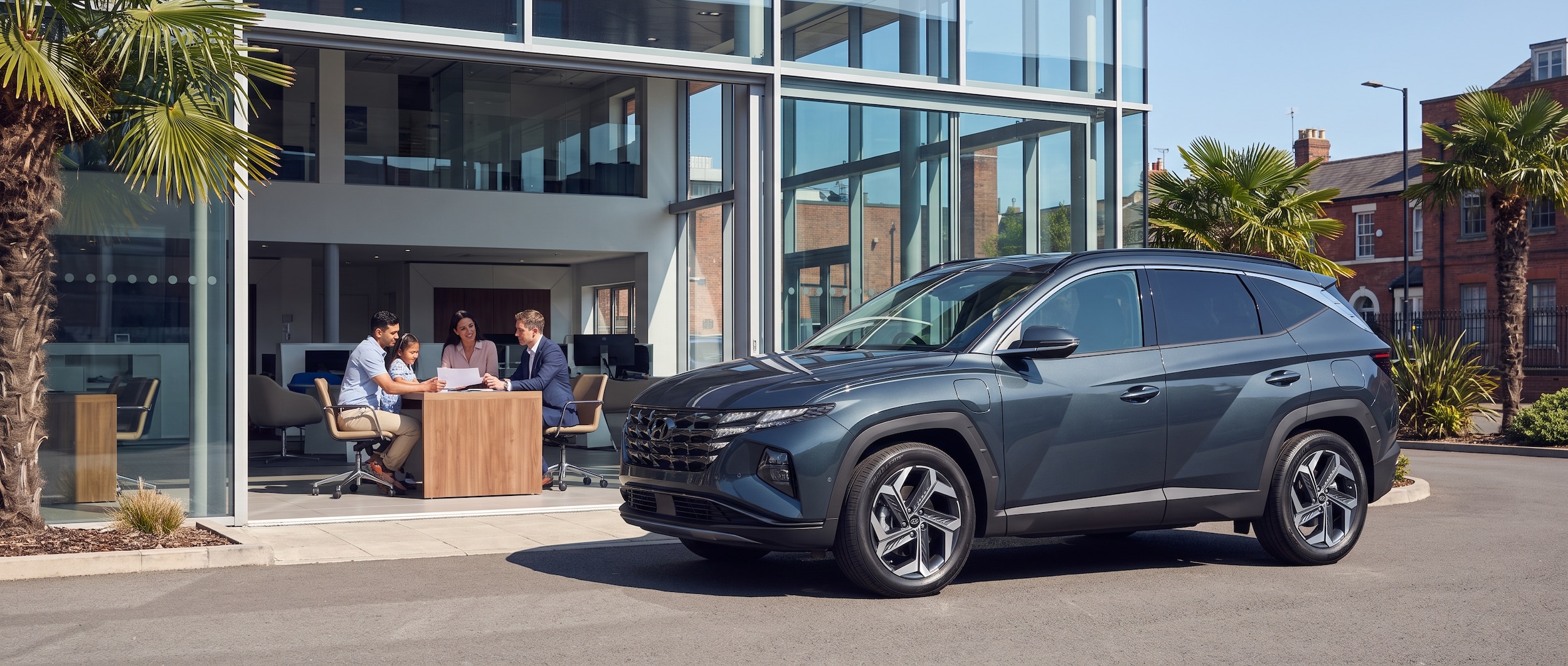 A 2026 Hyundai Tucson Hybrid parked in front of a modern dealership showroom on a sunny Birmingham summer day, with a family reviewing paperwork at a sales desk visible through the glass in the background