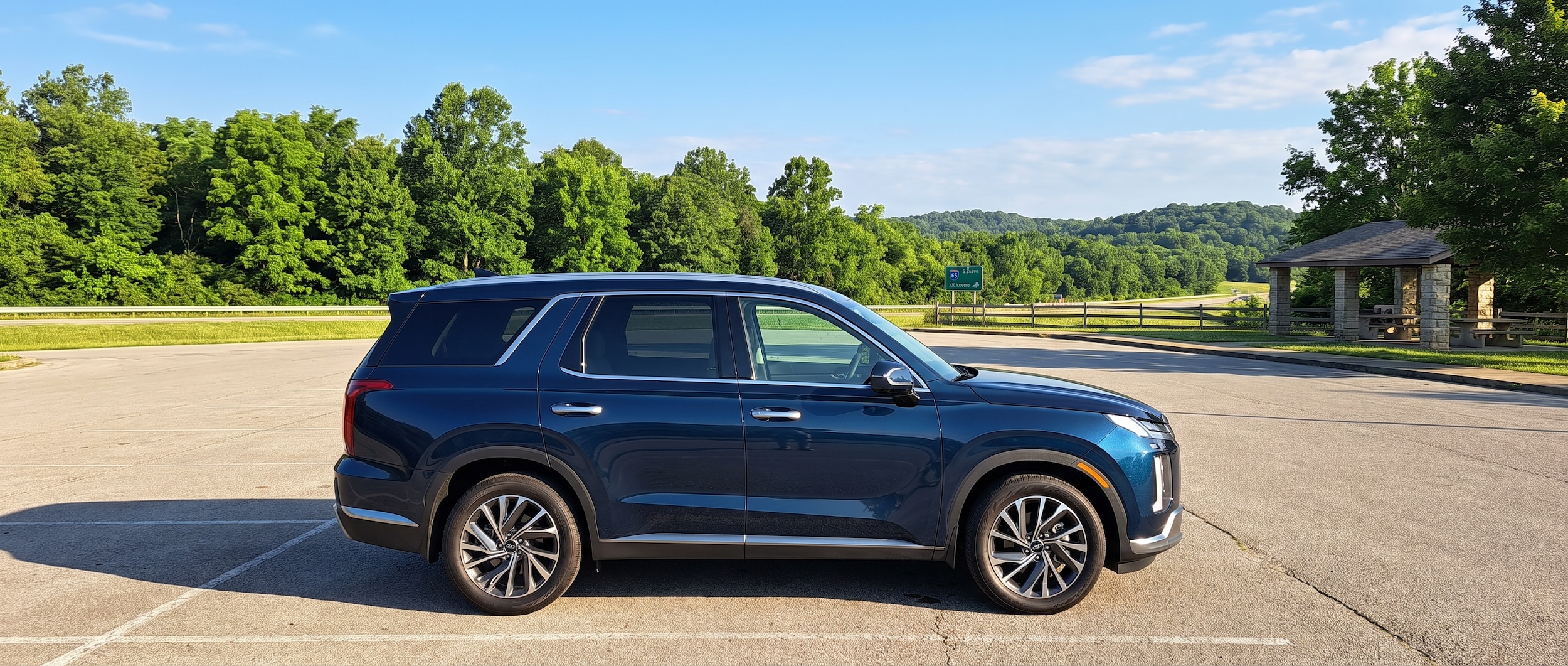 A 2026 Hyundai Palisade Hybrid in a deep metallic color loaded with gear, parked at a scenic Alabama rest stop along I-65 South on a sunny summer morning, with green tree lines and open sky in the background