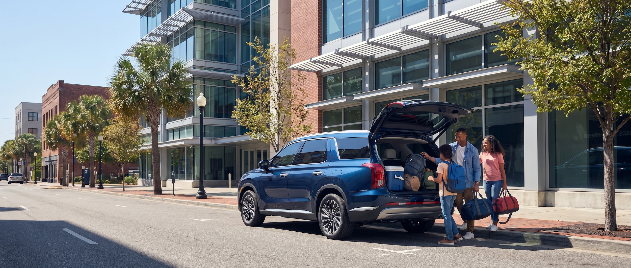 A 2026 Hyundai Palisade Hybrid in deep blue parked on a sunny downtown Birmingham street near a modern science museum building, family visible loading gear from the rear cargo area, summer daylight