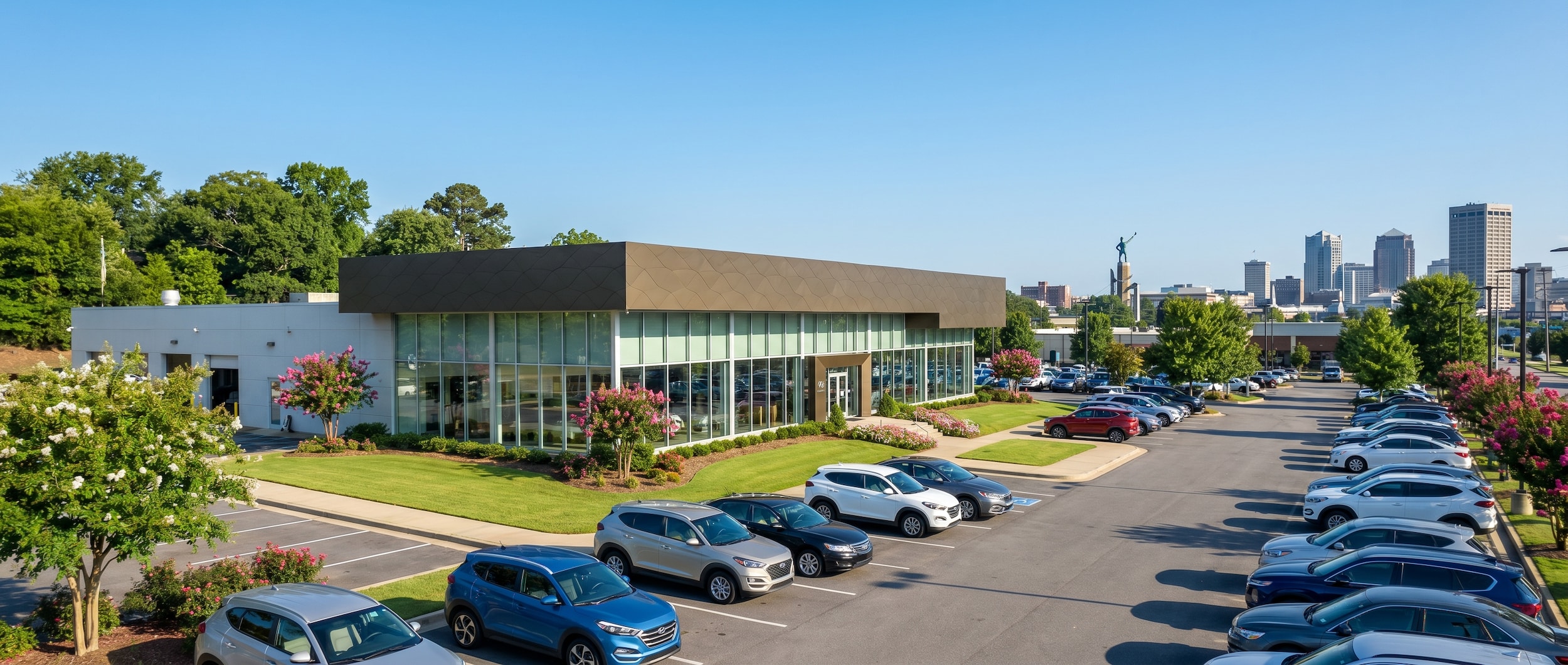 A bright, sunny afternoon view of a well-maintained Hyundai dealership exterior with a welcoming parking lot, green landscaping, and the Birmingham, Alabama skyline visible in the background distance.