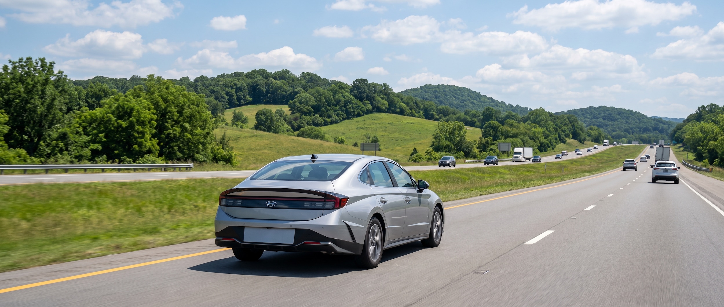 A 2026 Hyundai Sonata in silver or white cruising northbound on a wide Alabama interstate highway on a bright summer day with light cloud cover, green rolling hills visible in the background, representing the I-65 corridor between Birmingham and Huntsville in July