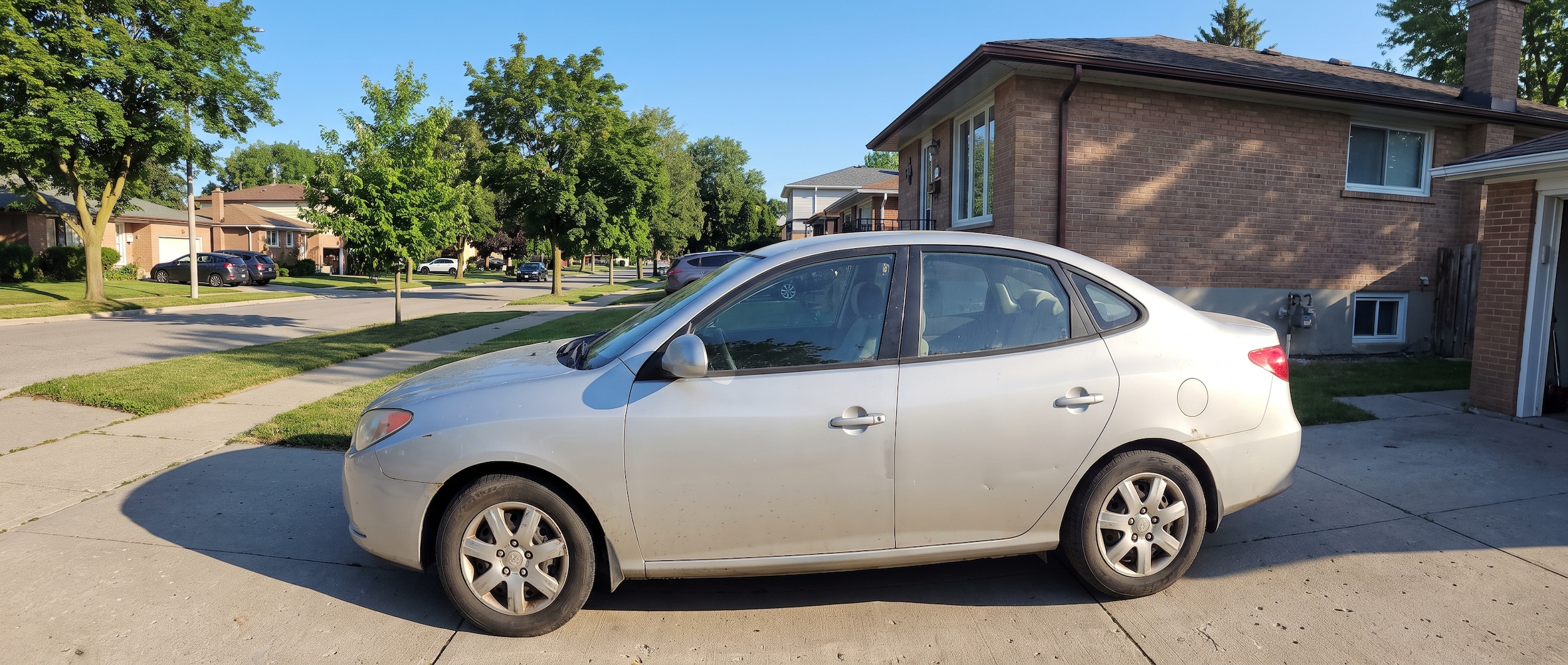 A silver used Hyundai Elantra parked in a suburban Tennessee driveway on a clear summer morning, with a tree-lined residential street in the background suggesting a Nashville suburb commuter neighborhood