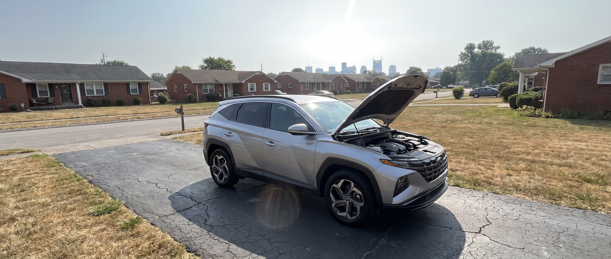 A 2026 Hyundai Tucson Hybrid parked in a sun-baked Tennessee suburban driveway on a bright July afternoon, hood partially raised, with the hazy Nashville skyline visible in the distance and heat shimmering off the asphalt