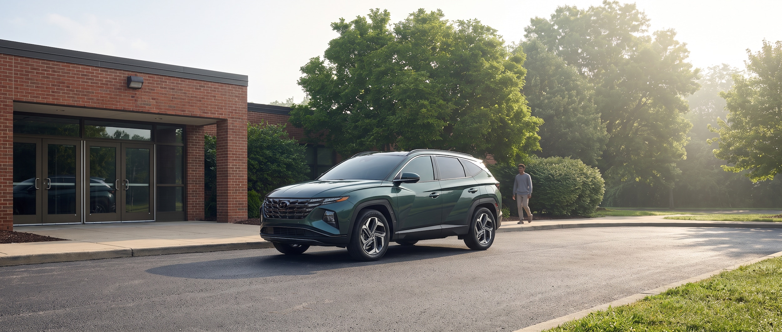 A 2026 Hyundai Tucson Hybrid in a suburban Tennessee setting, parked in front of a school entrance on a hot August morning, backpacks visible in the rear cargo area, green foliage in the background suggesting late summer