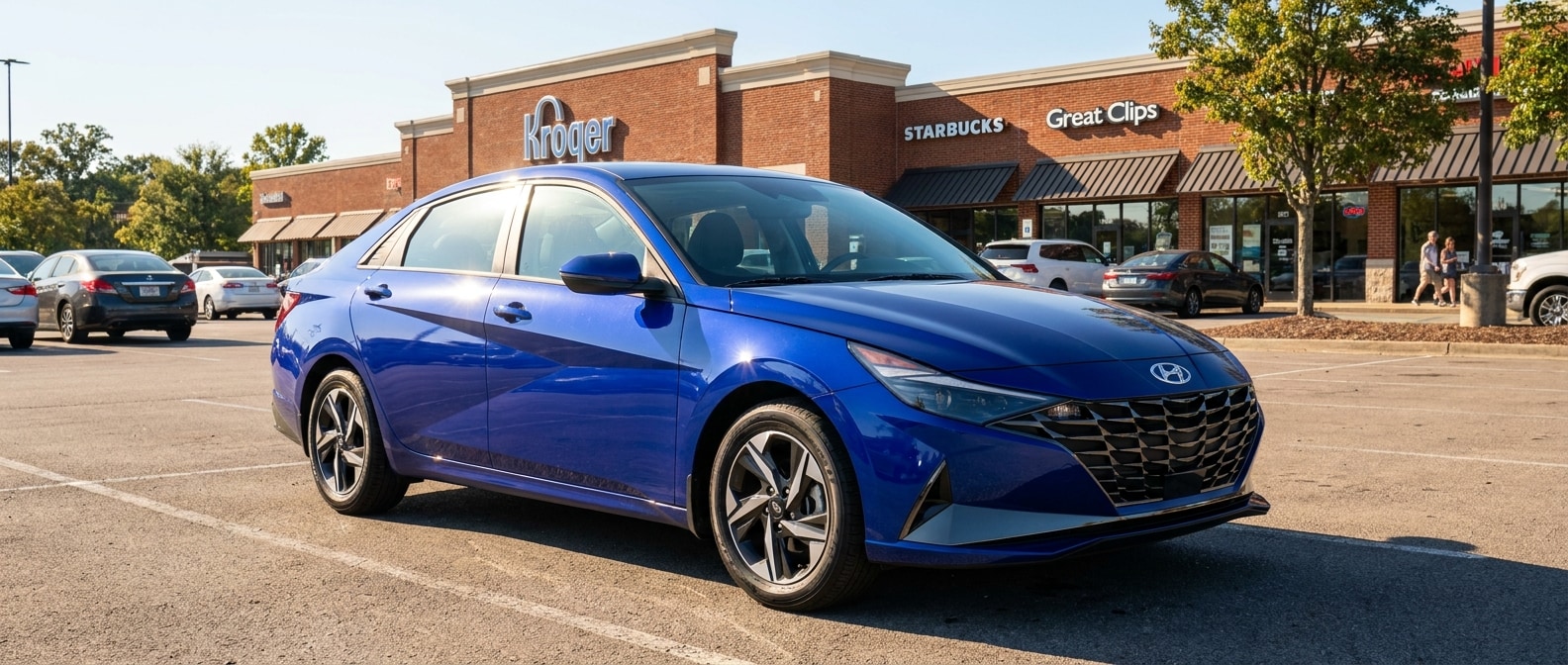 A 2026 Hyundai Elantra SE in Intense Blue parked in a suburban Tennessee shopping corridor on a bright summer afternoon, with a commercial retail strip visible in the background and sunlight reflecting off the car's angular parametric body lines