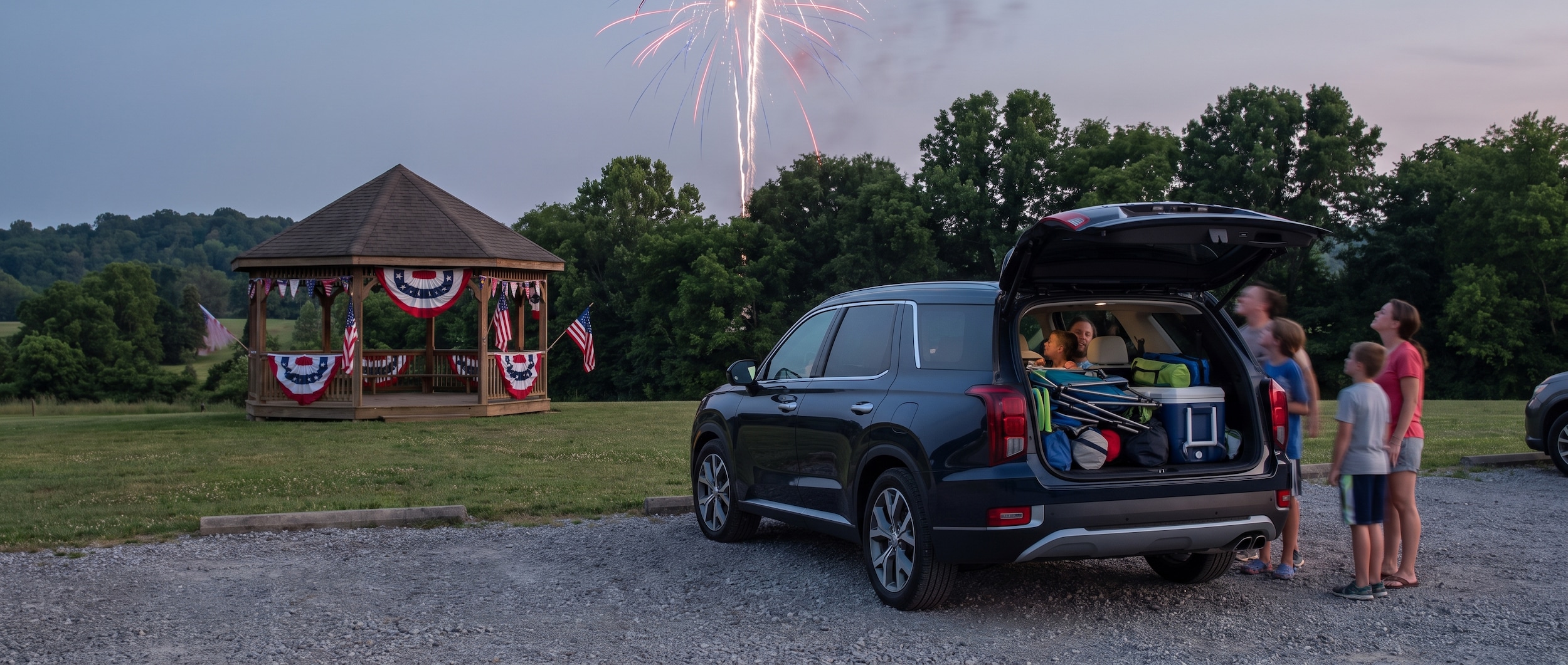 A dark-blue Hyundai Palisade loaded with passengers and a packed cargo area, parked at a community park in Middle Tennessee on a clear summer evening, with red-white-and-blue decorations visible in the background and fireworks beginning to light the sky overhead.