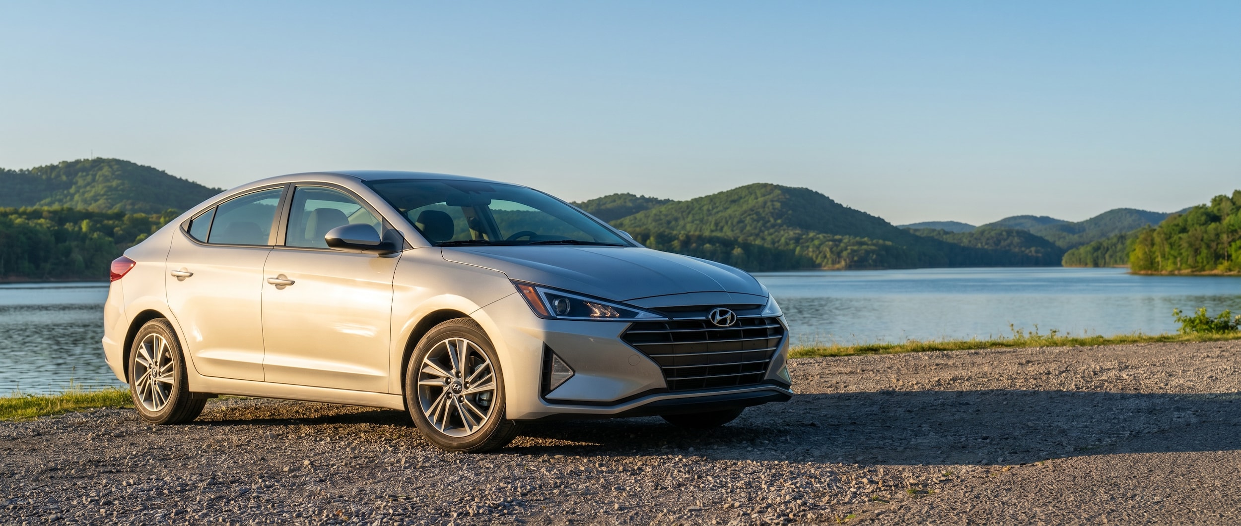 A silver Hyundai Elantra parked near a scenic Tennessee lake at golden hour, with rolling green hills visible in the background and a clear blue sky overhead.