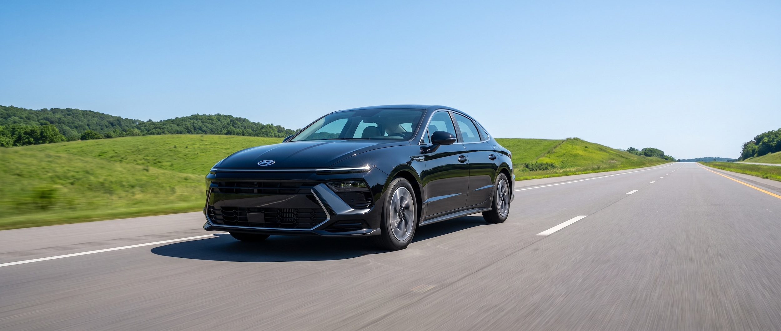 A 2026 Hyundai Sonata Hybrid sedan in Abyss Black cruising on a wide Tennessee interstate flanked by green rolling summer hills, low three-quarter front angle, clear blue sky, highway horizon ahead