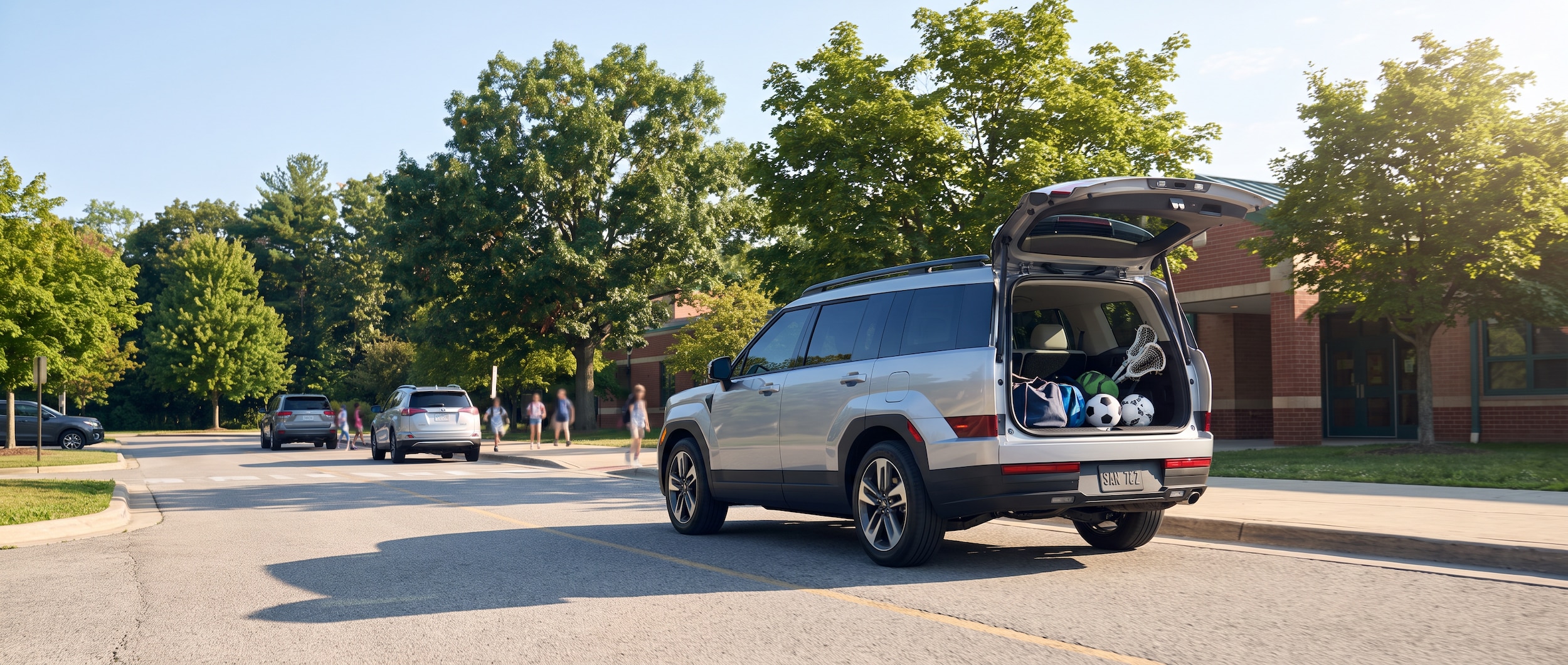A 2026 Hyundai Santa Fe in a silver or white exterior, shown pulling into a suburban school drop-off lane on a bright August morning in Tennessee, with a tree-lined school entrance in the background and a clear blue sky overhead. The SUV's hands-free liftgate is visible from a three-quarter rear angle, suggesting loaded cargo for fall sports.