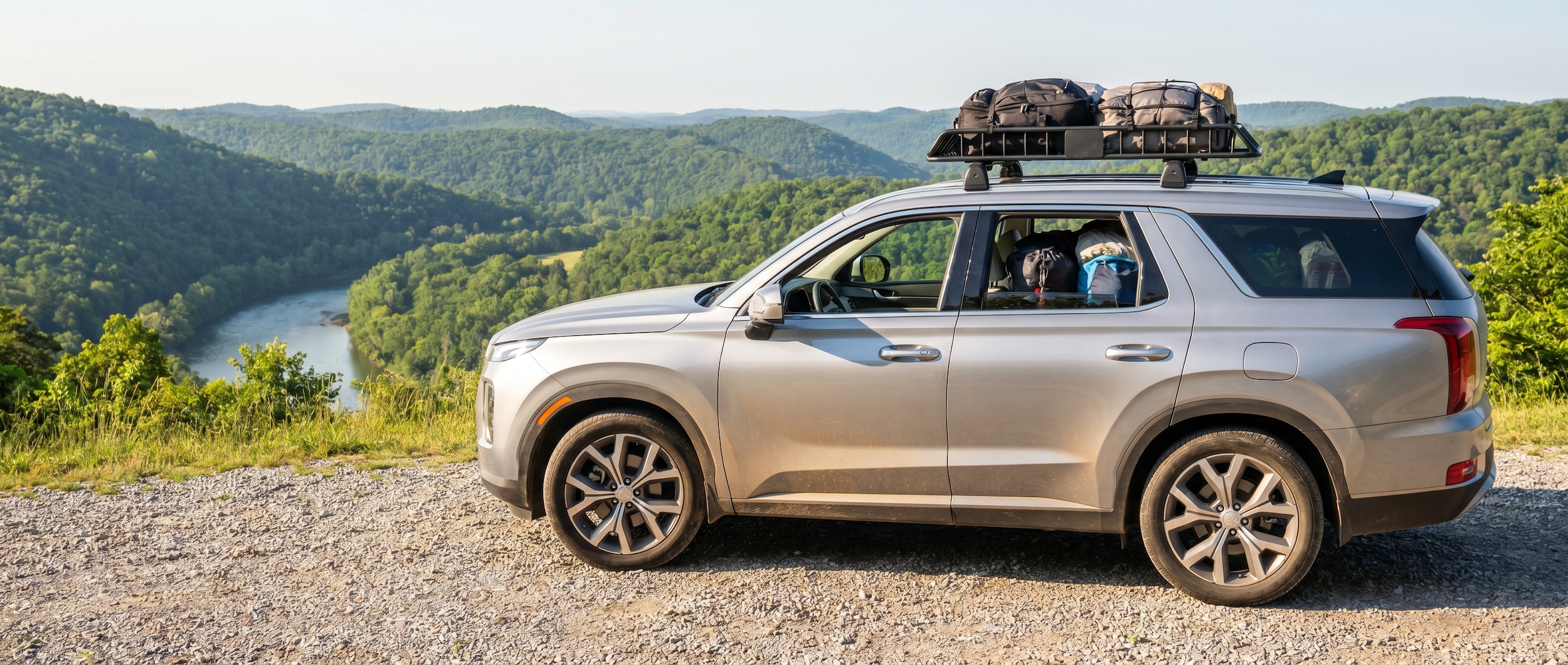 A silver Hyundai Palisade loaded for a road trip, parked on a scenic overlook with rolling Tennessee hills in the background on a sunny summer morning, luggage visible through the rear windows.