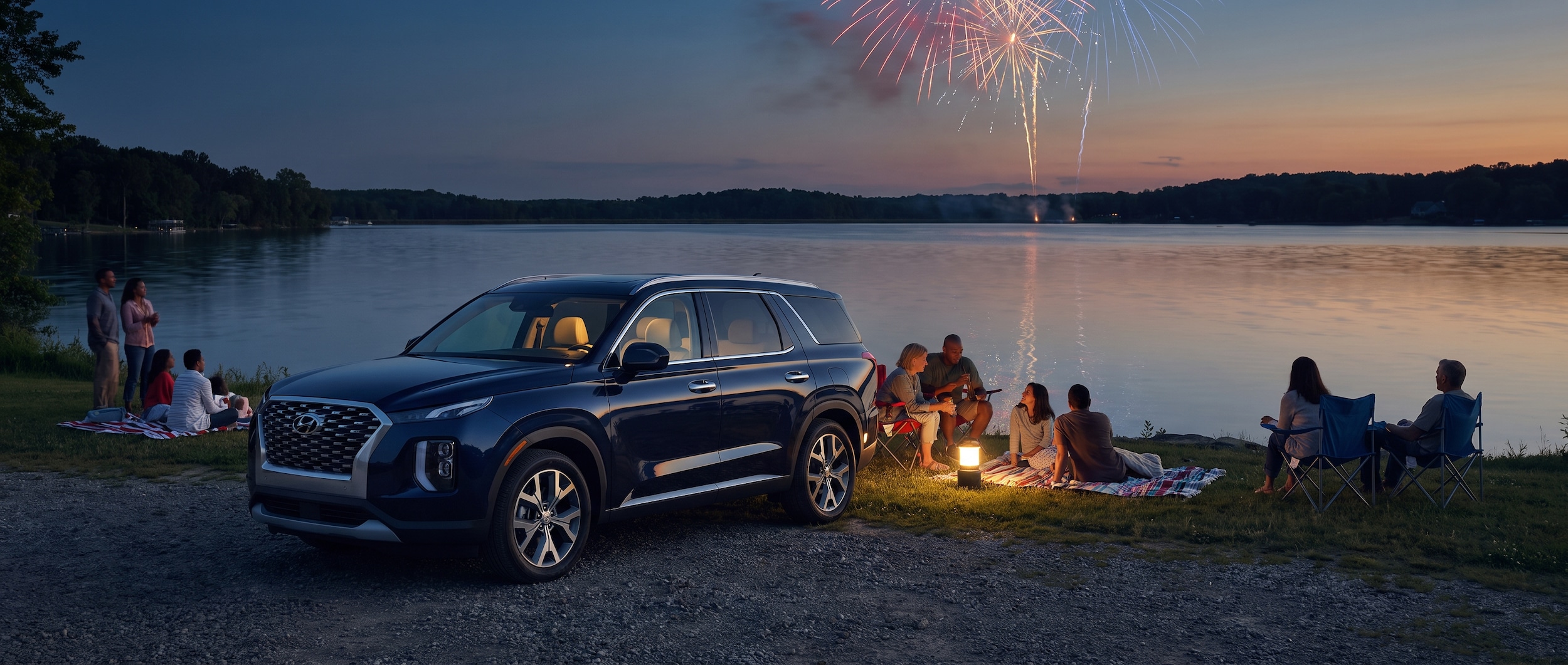 A spacious Hyundai Palisade parked near a lakeside area at dusk, with families setting up lawn chairs and the glow of fireworks beginning to light up the sky over the water