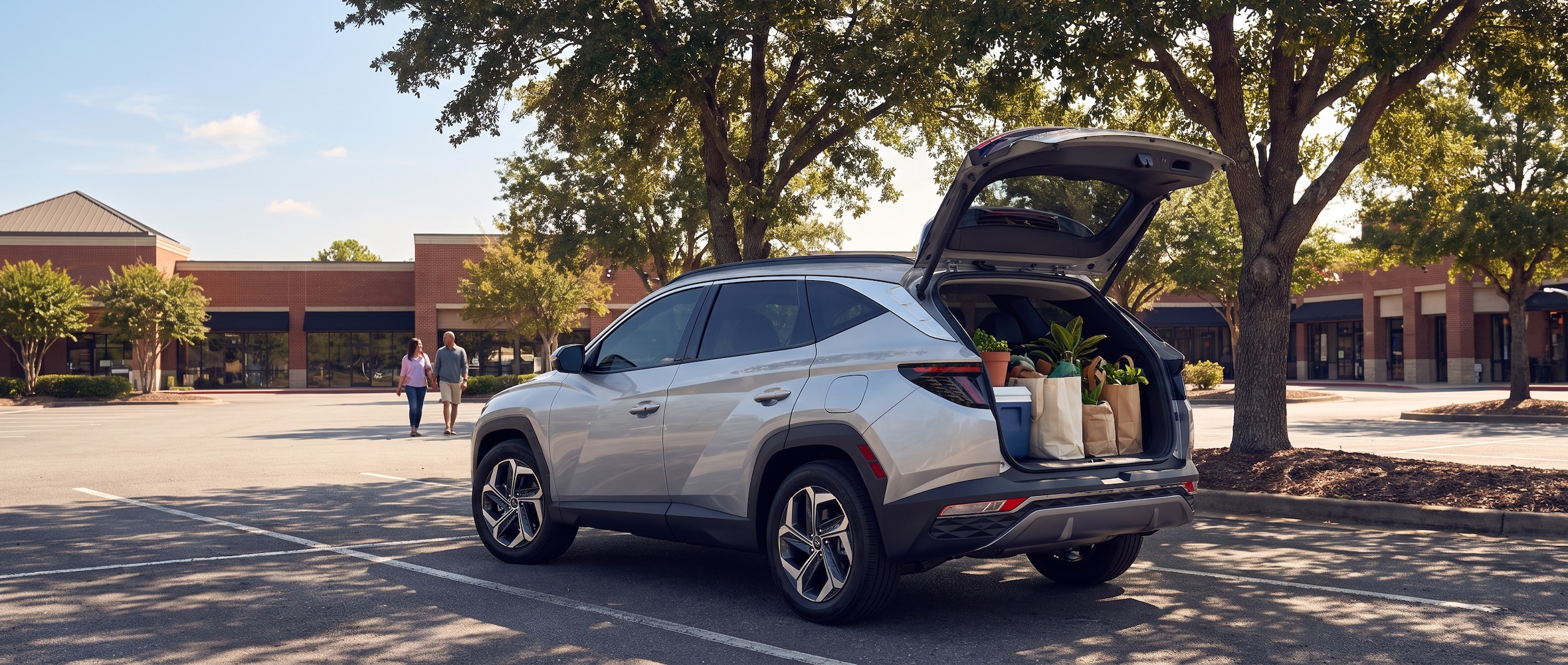 A silver or white Hyundai Tucson Hybrid parked in an open-air lifestyle shopping center lot on a bright Tennessee summer day, mature trees providing partial shade and a warm blue sky overhead, the SUV's liftgate open revealing loaded shopping bags suggesting a productive summer errand run