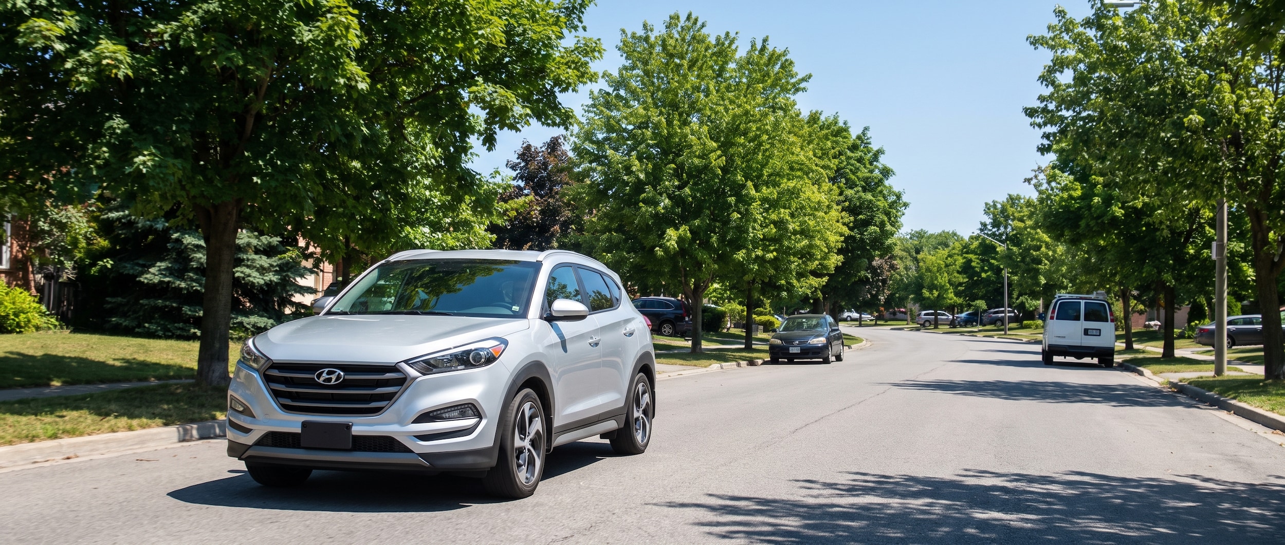 A silver Hyundai Tucson driving along a sun-drenched suburban road lined with green summer trees, with a clear blue sky overhead and light traffic visible in the background.
