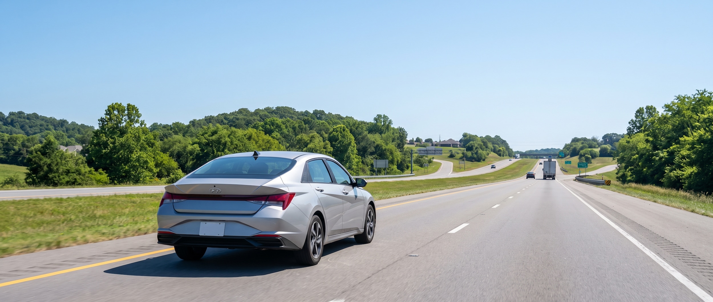 A 2026 Hyundai Elantra in silver or blue driving on a sunny Tennessee four-lane controlled-access highway, mid-morning summer light, light traffic visible in the distance, trees and suburban exits on either side, clear sky