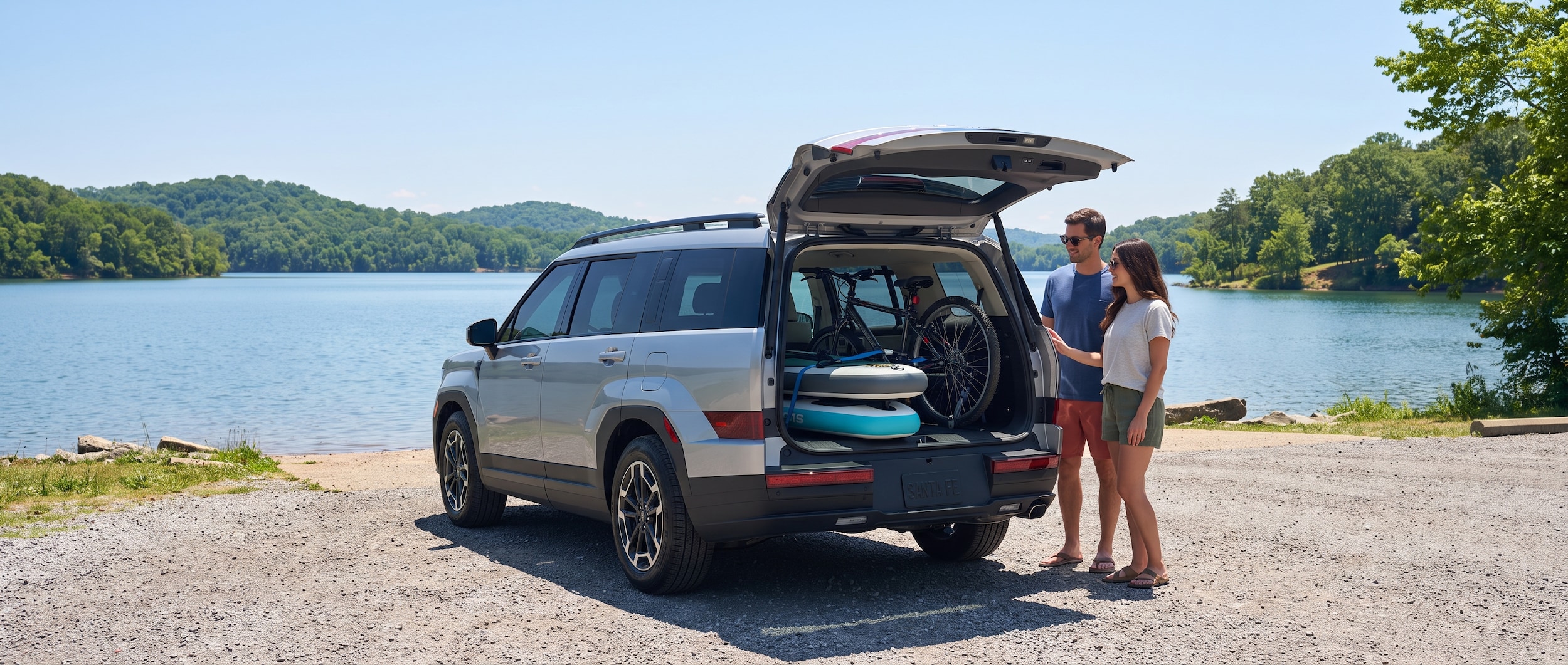 A 2026 Hyundai Santa Fe with its liftgate open, loaded with paddleboards and a bicycle, parked at a Tennessee lake access point on a bright summer day, showing the wide, flat cargo floor