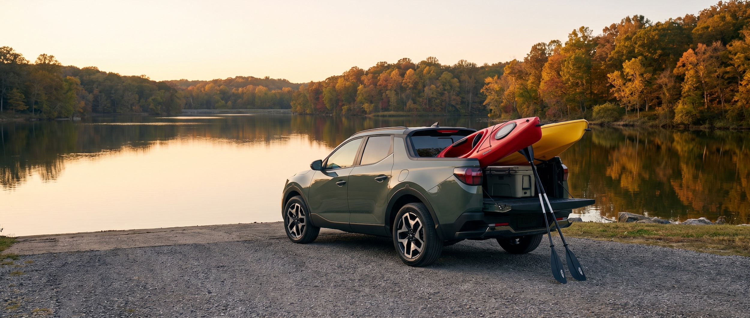 A Hyundai Santa Cruz parked near a lake boat ramp at golden hour with kayaks loaded in the truck bed, paddles resting against the tailgate, and a cooler visible in the cargo area, surrounded by Tennessee trees reflecting on calm water.