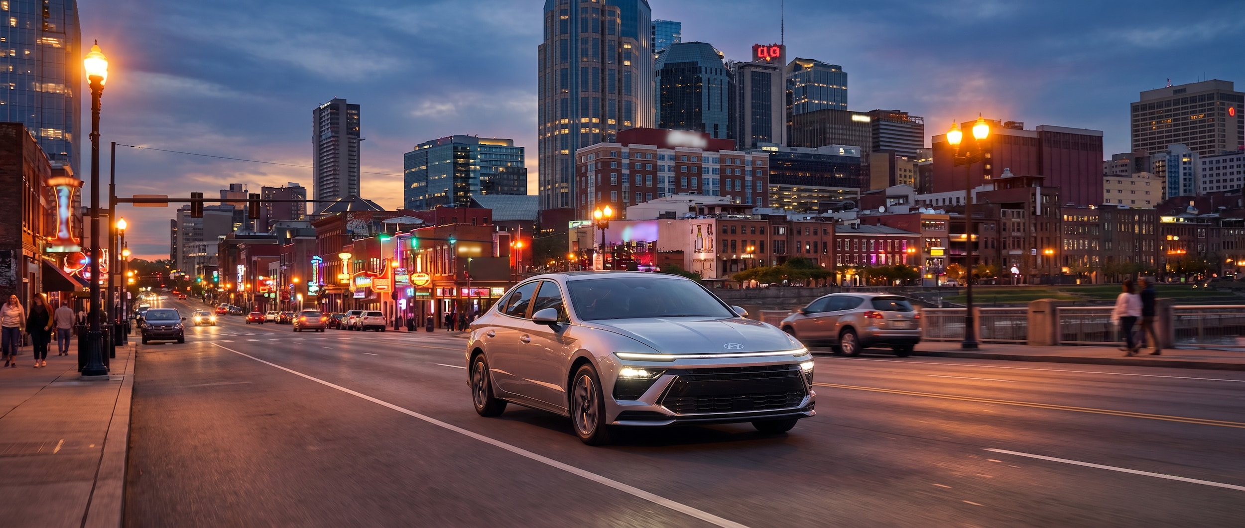 A 2026 Hyundai Sonata Hybrid in a silver or deep blue finish driving along a city boulevard at dusk, downtown Nashville skyline with lit-up honky-tonk neon signs visible in the background, warm summer evening atmosphere