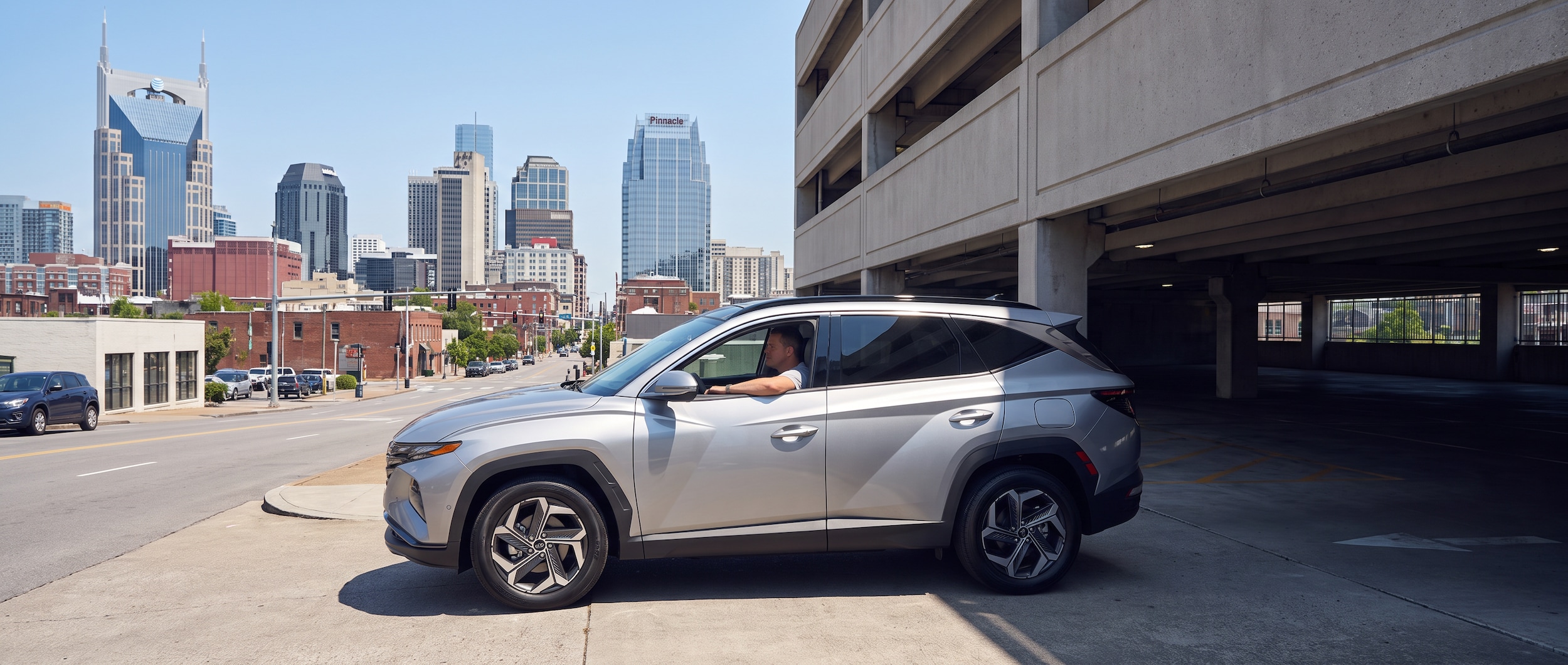 A 2026 Hyundai Tucson Hybrid in silver pulling into a downtown Nashville parking garage entrance, with the city's skyline visible in the background on a sunny summer day