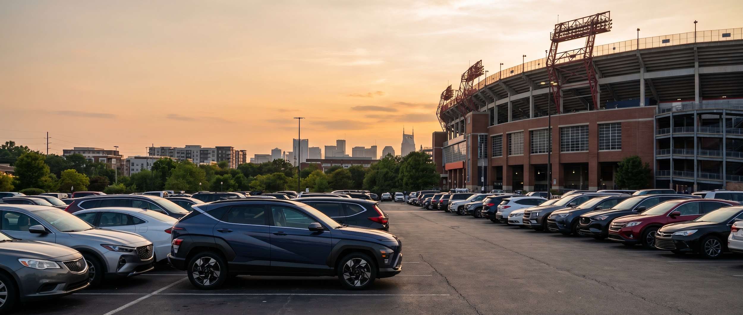 A 2026 Hyundai Kona in a compact stadium parking lot near a large sports venue, shot from a slightly elevated angle on a sunny summer evening, Nashville skyline faintly visible in the background.
