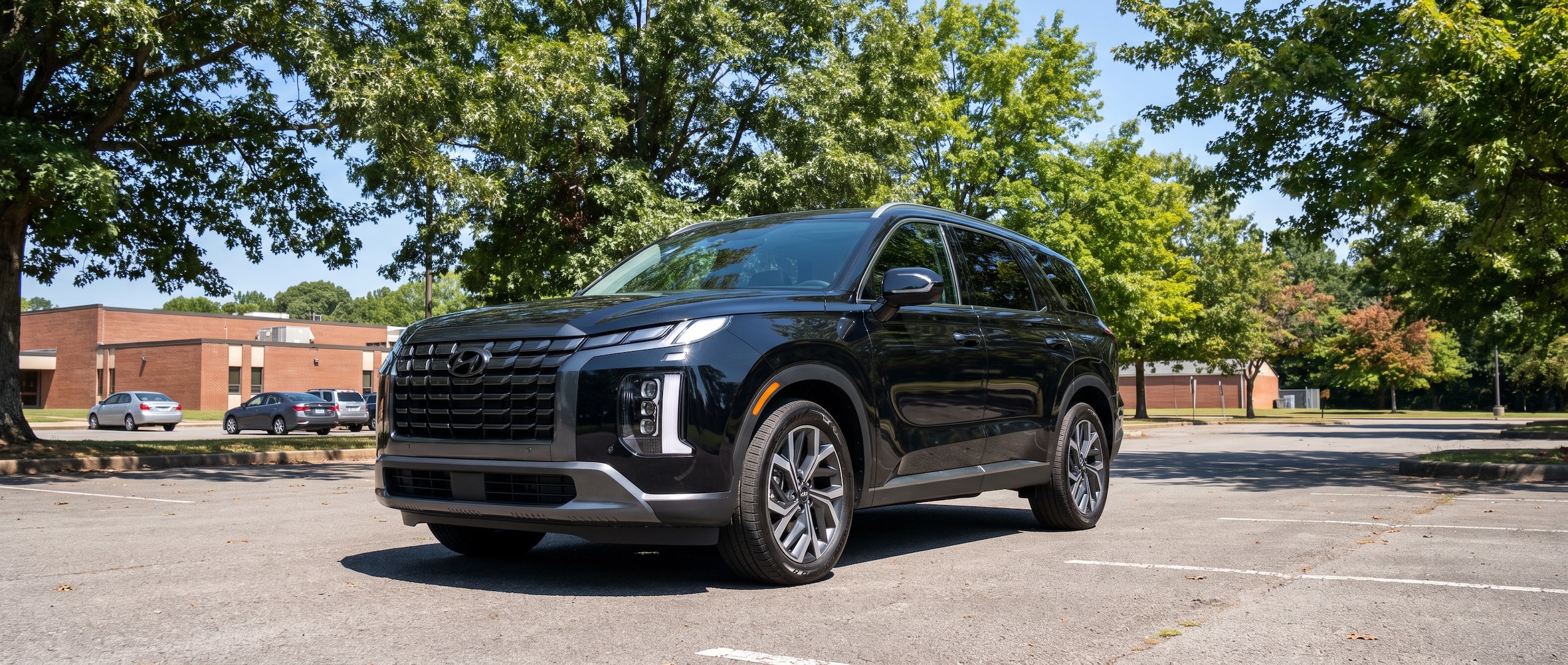 A 2026 Hyundai Palisade in Abyss Black parked in a suburban Tennessee school parking lot in summer, with green tree canopy overhead and a clear blue sky, shot from a three-quarter front angle showing the bold vertical LED grille and wider stance
