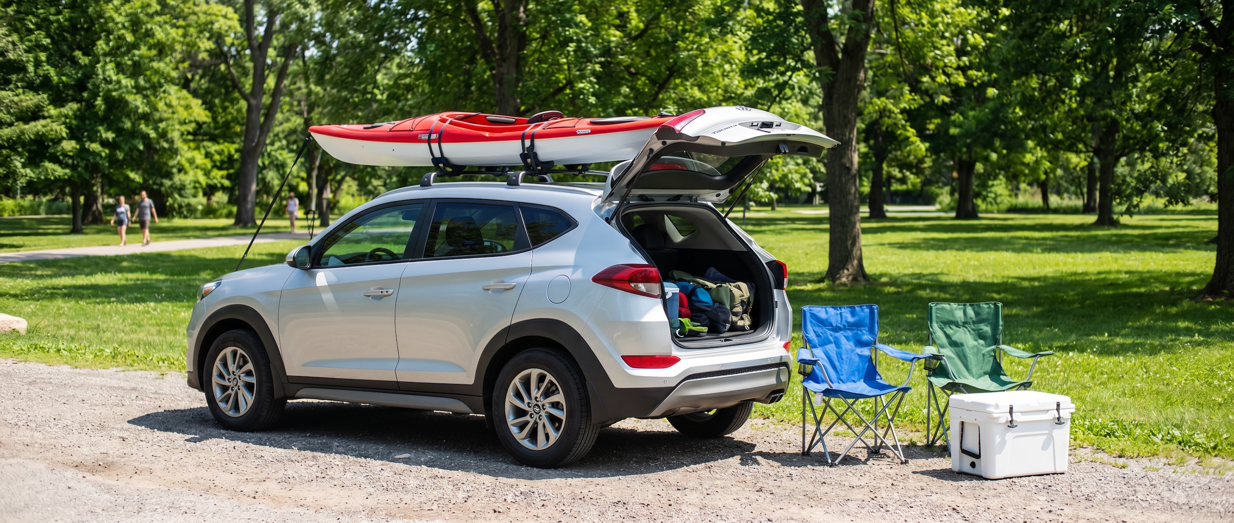 A silver Hyundai Tucson parked near a green park setting on a sunny summer day, with a kayak, folding chairs, and a cooler visible beside the open rear hatch, suggesting an outdoor adventure outing