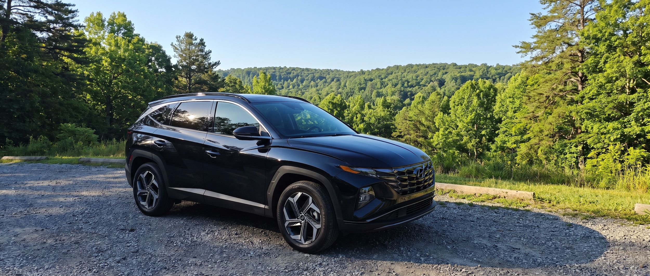 A 2026 Hyundai Tucson Hybrid in Abyss Black parked at a Tennessee state park trailhead on a bright summer morning, green forested plateau in the background, gear visible through the rear window, blue sky overhead