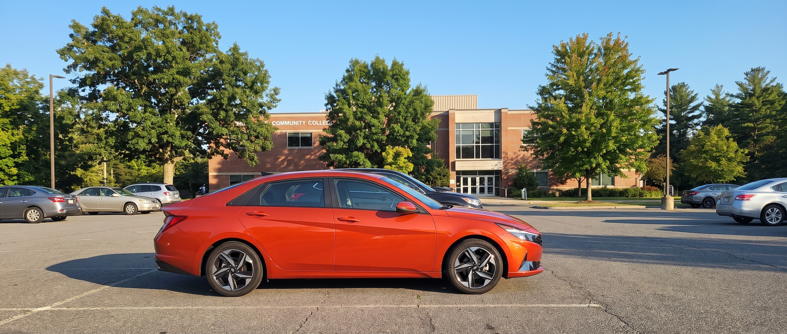 A 2026 Hyundai Elantra in a bright compact color parked on a sunny suburban Tennessee campus parking lot, with a community college building and green trees in the background, evoking a back-to-school August mood.