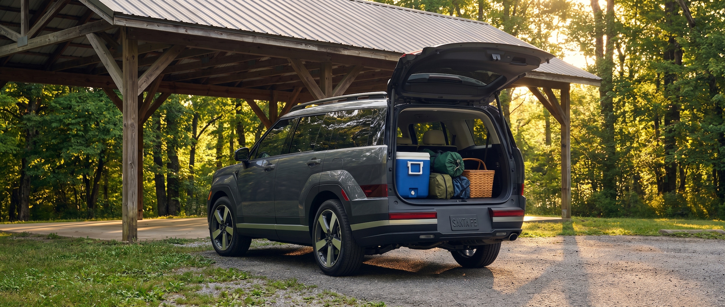 A 2026 Hyundai Santa Fe in Ecotronic Gray parked at a shaded park pavilion in summer, liftgate open revealing a full cargo area with a large cooler, folding camp chairs, and a picnic bag, lush Tennessee trees in the background, warm afternoon light