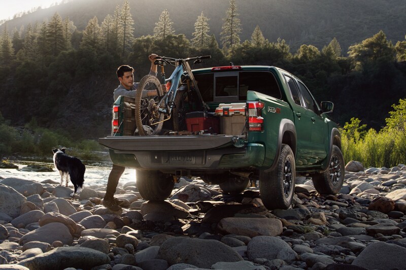 Still of a man loading his bike into the bed of a 2023 Nissan Frontier by a riverbed with his dog nearby.png