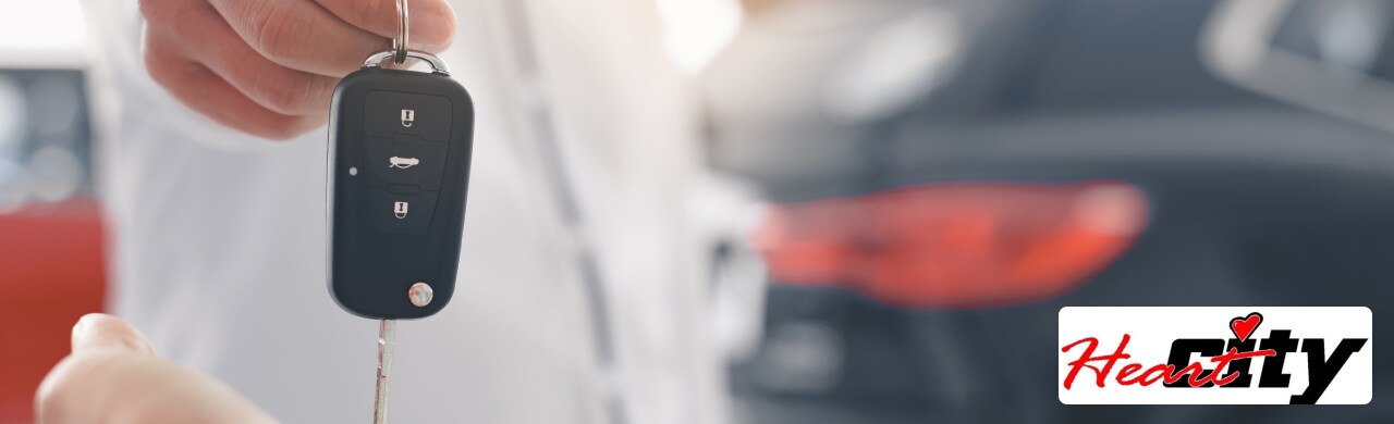A hand is handing over a car key with a silver car charm on the keychain.