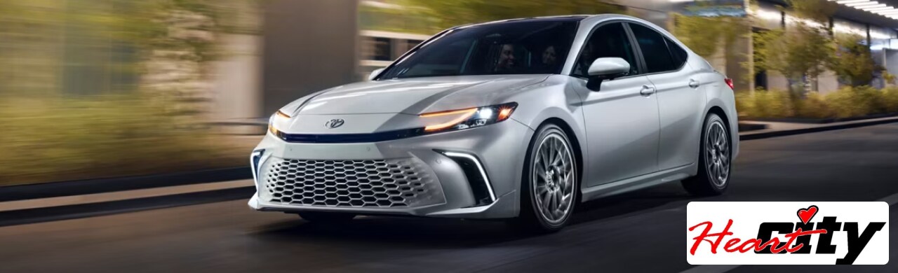 The front view of a sleek white 2024 Toyota Camry with stylish headlights, driving on a city street.