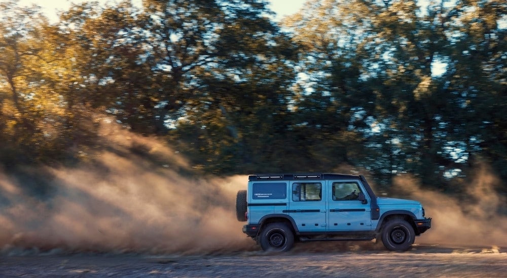 A white 2025 INEOS Grenadier Station Wagon is shown kicking up dust off-road.