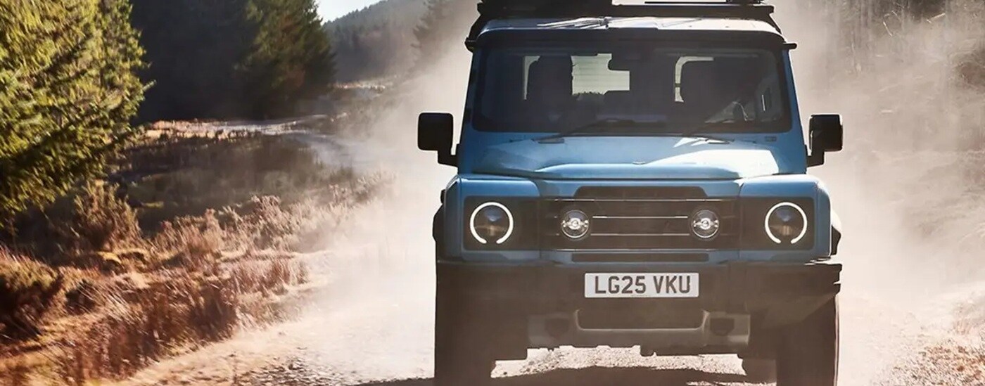 Front of a blue 2025 INEOS Grenadier driving off-road down a dirt road.