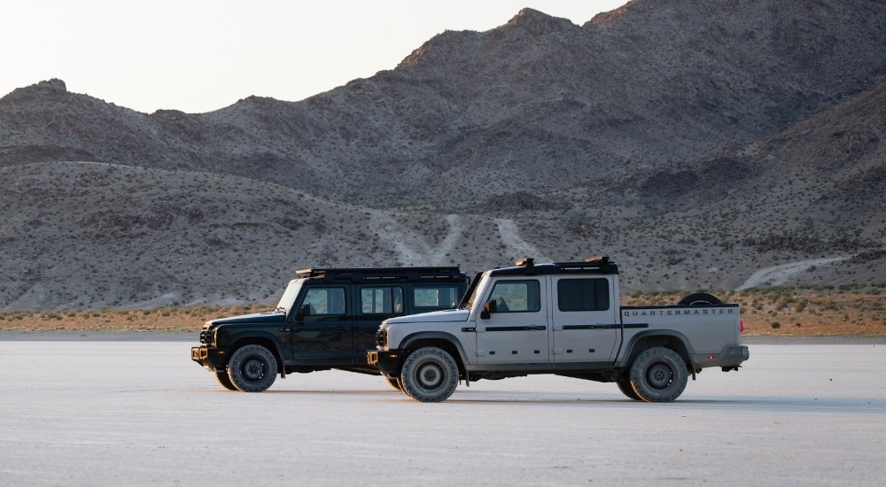 A black 2025 INEOS Grenadier Station Wagon and white Quartermaster are parked off-road.