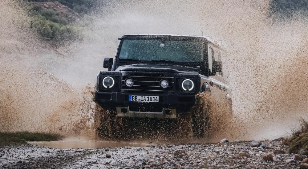 A black 2025 INEOS Grenadier Station Wagon is splashing through a puddle after leaving a INEOS dealer.