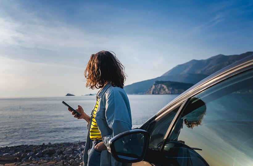 Woman leaning against a car while on the phone and looking out over the ocean