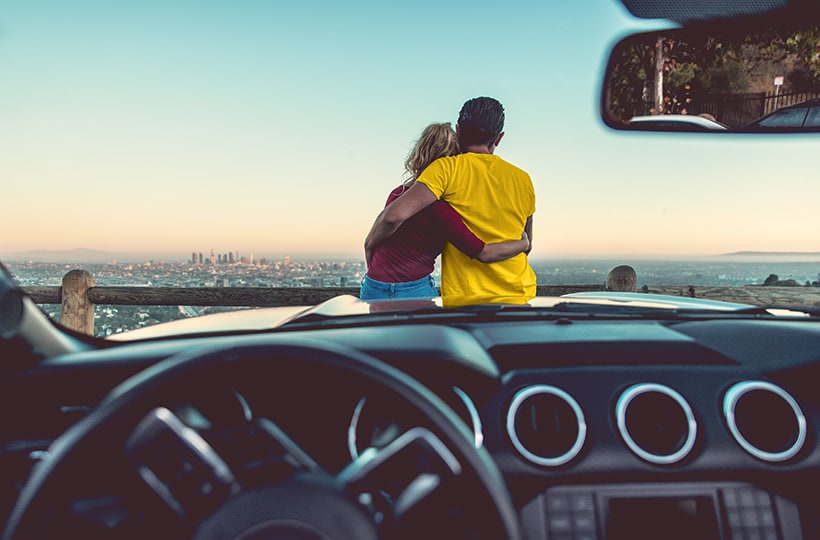 A couple hugging while sitting on hood of a car and looking out over city skyline