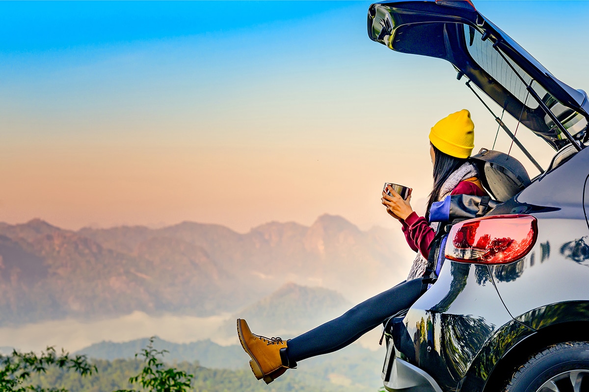 A woman sitting on rear bumper of car with rear hatch open while looking out over a mountain view