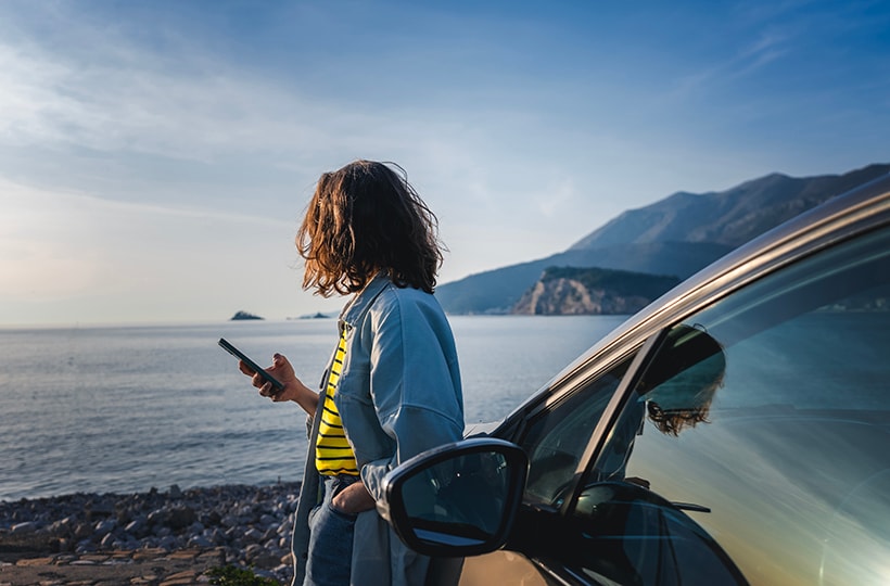 A woman using a phone while leaning on her car and looking out over a body of water